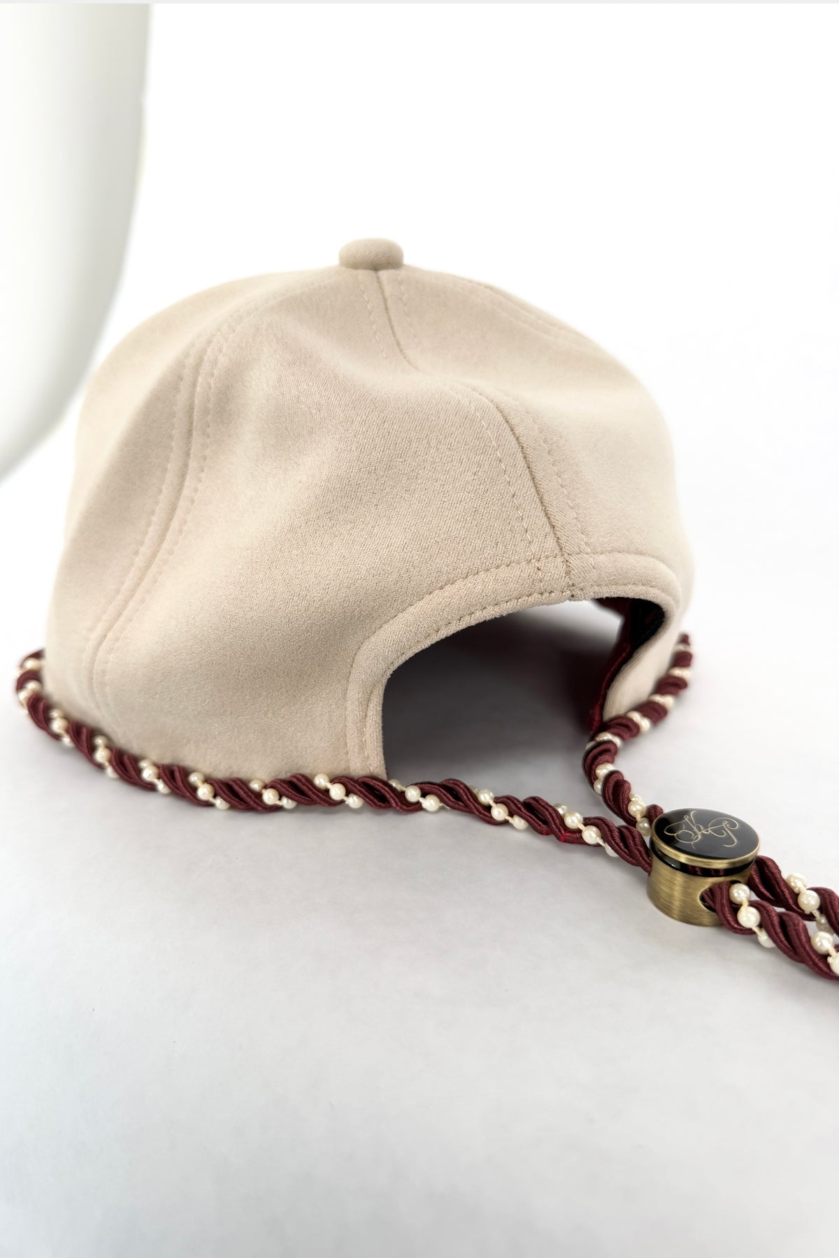 A ivory aviator-style cap with a braided brown and cream cord and a small gold-toned round charm, photographed on a white studio background.