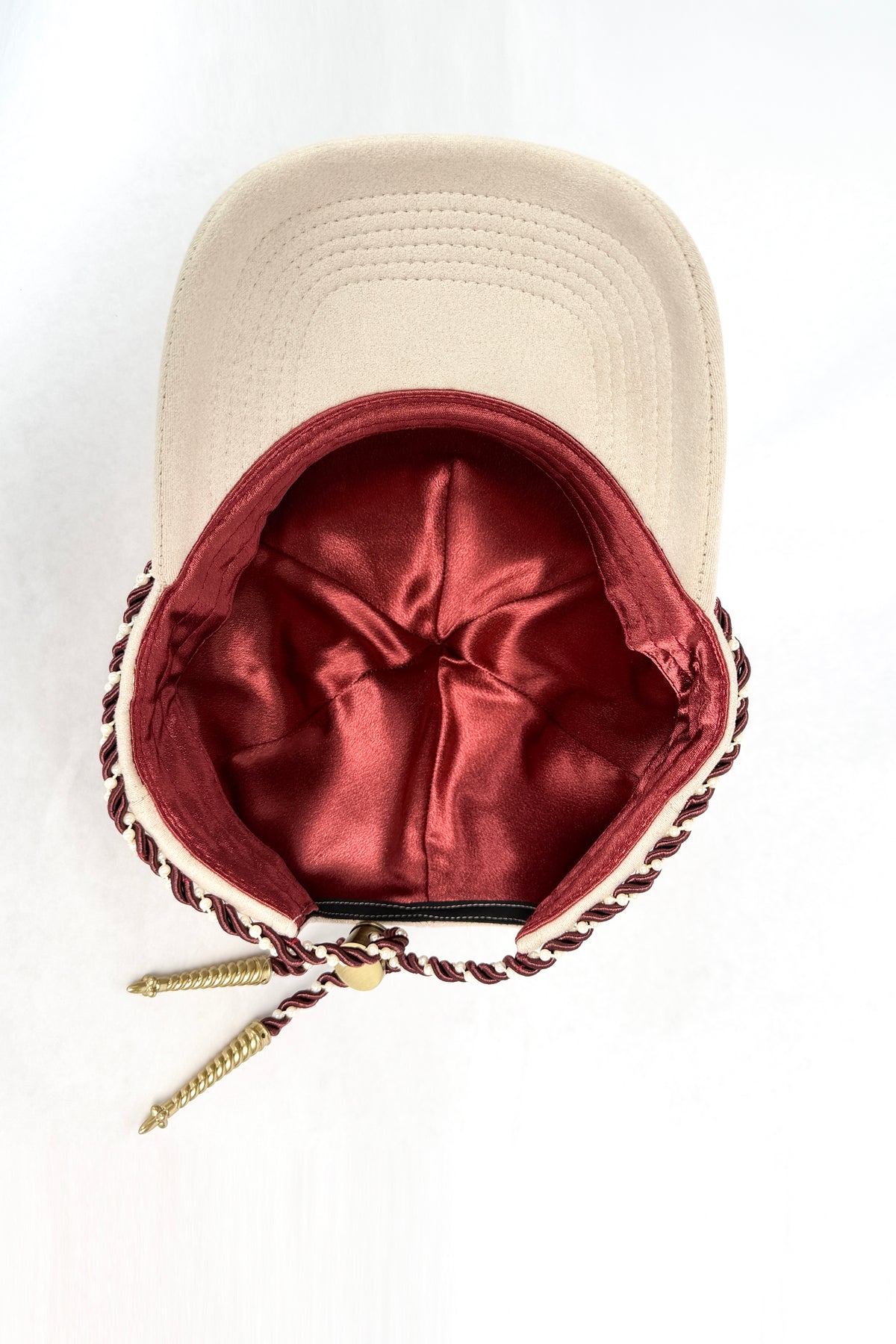 An ivory-colored baseball cap with a deep red satin-lined interior and a decorative braided red and gold cord detail around the edge.