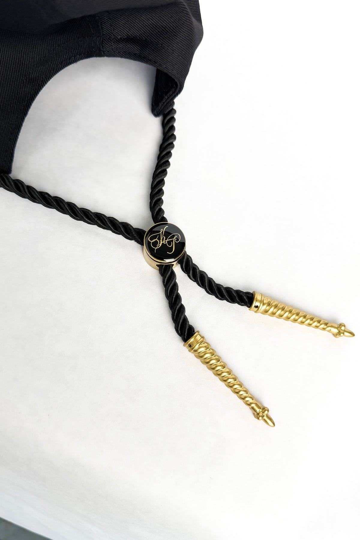 A close-up of a black twisted rope bolo tie with gold-toned decorative ends and a central round black medallion featuring gold script, displayed against a white background with a black fabric accessory partially visible at the top.