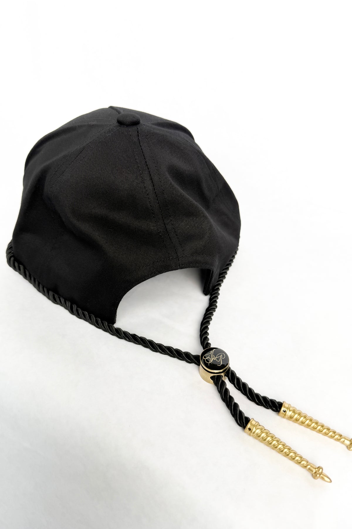 A black fabric cap with braided black ties and gold-toned decorative end pieces, photographed on a plain white background.