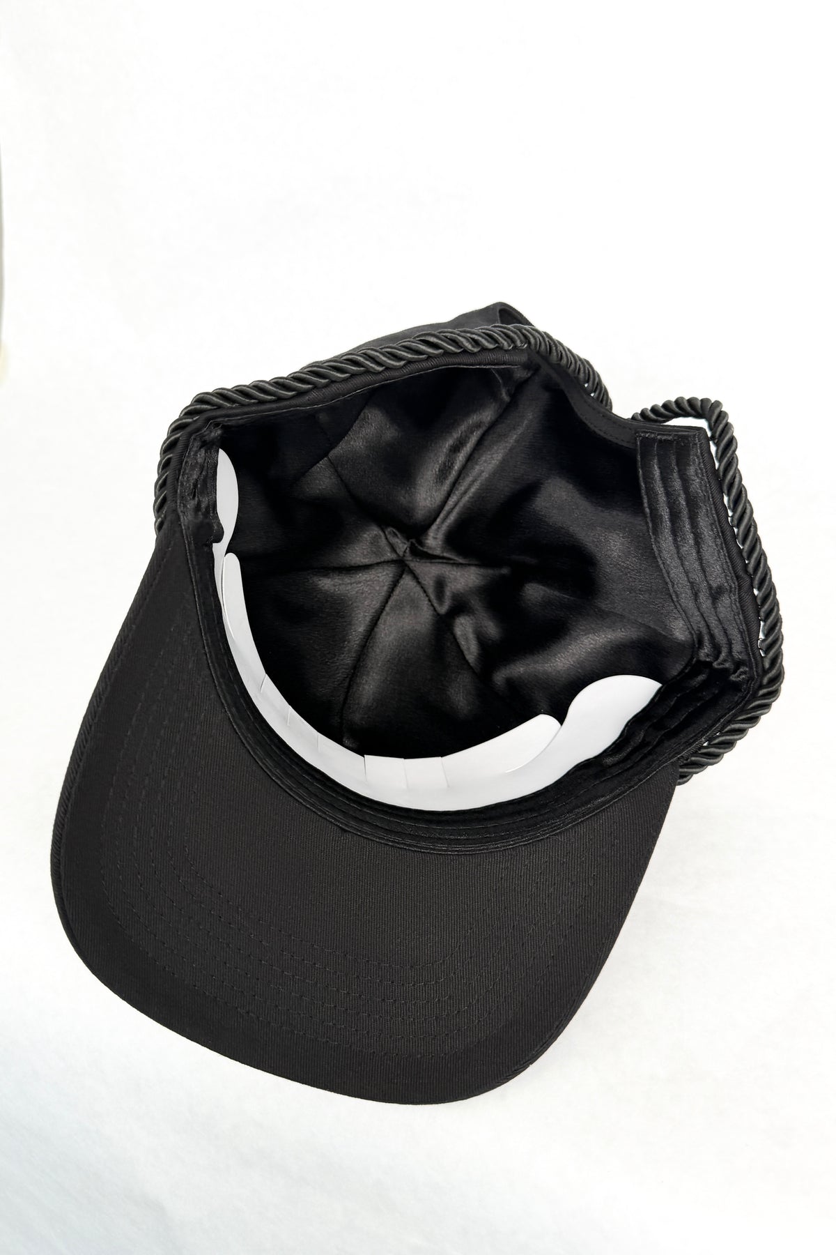 Black baseball cap with a black satin inner lining and a black braided rope detail along the top edge, shown from a top-down angle on a white background.