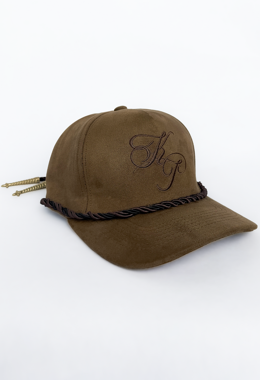 A brown moleskin baseball cap with a dark brown braided rope detail wrapped around the brim and a matching embroidered monogram logo on the front. The cap is displayed on a clean white background with a subtle shadow.