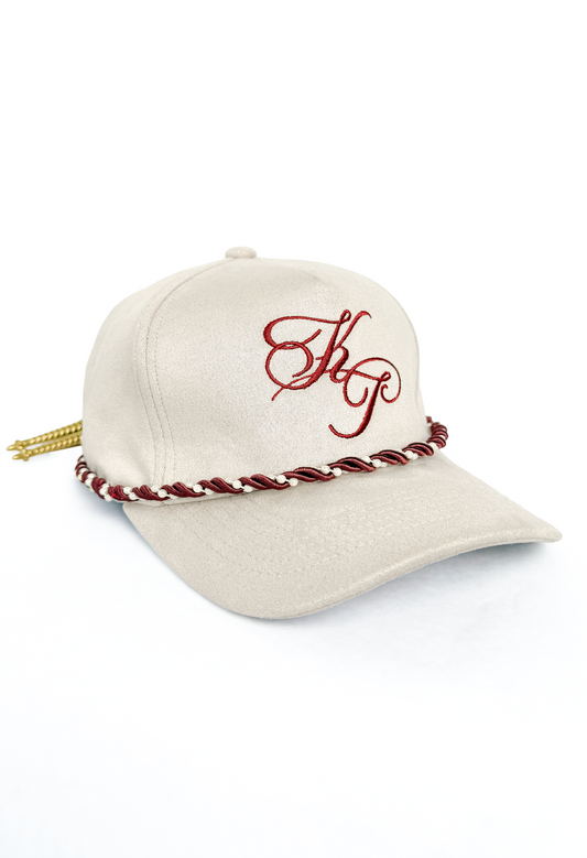 An ivory baseball cap featuring a red embroidered monogram on the front, a red and white braided rope detail around the base of the crown, and a gold tassel hanging from the side, set against a plain white background.