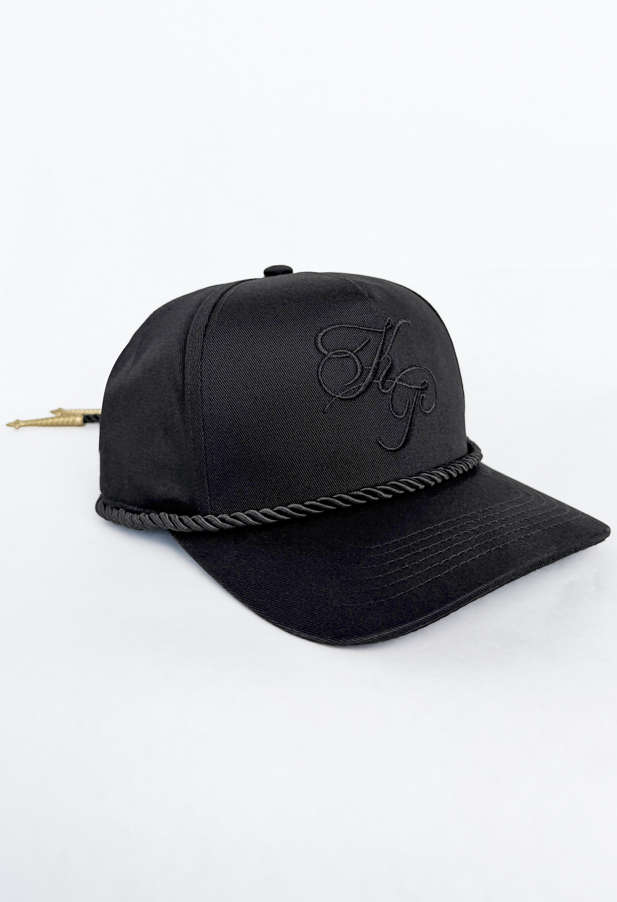 A black baseball cap featuring a black embroidered monogram logo on the front, a braided rope detail across the brim, and a small gold arrowhead accent on the side, set against a plain white background.