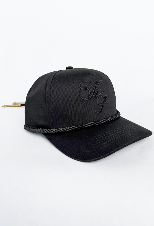 A black baseball cap featuring a black embroidered monogram logo on the front, a braided rope detail across the brim, and a small gold arrowhead accent on the side, set against a plain white background.